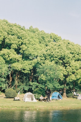 A serene outdoor scene with several tents pitched on a grassy area adjacent to a body of water. Lush green trees form a canopy in the background, providing shade and an inviting natural backdrop. People are seen engaging in leisure activities near the tents, enjoying a peaceful day in nature.