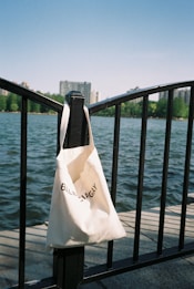 A white tote bag with black text hangs from a black metal railing overlooking a body of water. In the background, there are city buildings partially obscured by greenery, under a clear blue sky.