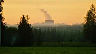 A countryside landscape at sunrise or sunset with a power plant emitting smoke in the distance. The scene includes lush green fields in the foreground, scattered trees, and a small soccer goalpost. A clear sky with soft orange hues creates a warm atmosphere.