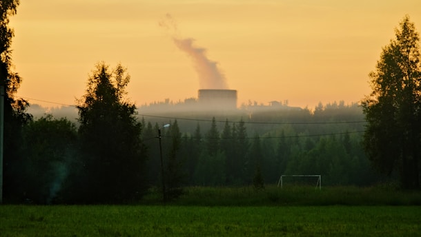 A countryside landscape at sunrise or sunset with a power plant emitting smoke in the distance. The scene includes lush green fields in the foreground, scattered trees, and a small soccer goalpost. A clear sky with soft orange hues creates a warm atmosphere.