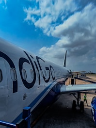 An airplane on the tarmac with the logo 'IndiGo' is parked, with a clear view of its fuselage and several windows along the side. The sky above is partly cloudy, and there is a boarding bridge connected to the aircraft. A person in a safety vest is visible near the aircraft's landing gear.