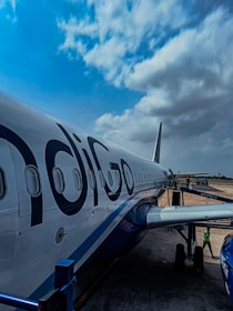 An airplane on the tarmac with the logo 'IndiGo' is parked, with a clear view of its fuselage and several windows along the side. The sky above is partly cloudy, and there is a boarding bridge connected to the aircraft. A person in a safety vest is visible near the aircraft's landing gear.