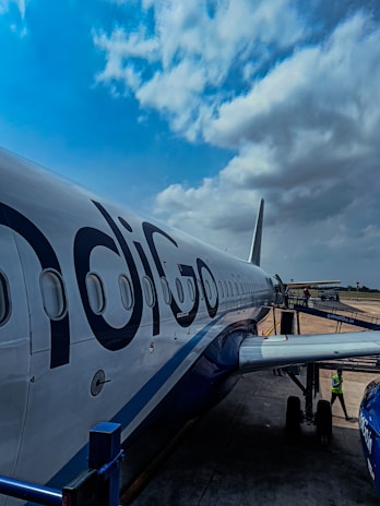 An airplane on the tarmac with the logo 'IndiGo' is parked, with a clear view of its fuselage and several windows along the side. The sky above is partly cloudy, and there is a boarding bridge connected to the aircraft. A person in a safety vest is visible near the aircraft's landing gear.