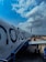 An airplane on the tarmac with the logo 'IndiGo' is parked, with a clear view of its fuselage and several windows along the side. The sky above is partly cloudy, and there is a boarding bridge connected to the aircraft. A person in a safety vest is visible near the aircraft's landing gear.