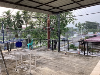 A covered balcony area overlooking a street. It has a metal railing and a few visible items like a turquoise chair, a blue piece of cloth on a drying rack, and a white metal rack. There are trees in the background and utility lines running across the view.