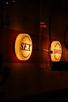 Illuminated circular signs with the words 'SEX' and 'DRUGS' are mounted on a wall. The signs emit a warm glow against a dimly lit background, creating a moody atmosphere.