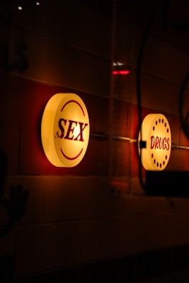 Illuminated circular signs with the words 'SEX' and 'DRUGS' are mounted on a wall. The signs emit a warm glow against a dimly lit background, creating a moody atmosphere.