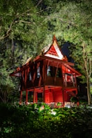An elevated traditional wooden house with a steeply pitched roof is illuminated at night. It is surrounded by lush greenery, with tall trees and various plants visible in the foreground. The house is painted red, with light highlighting its architectural details and casting shadows in the surrounding foliage.
