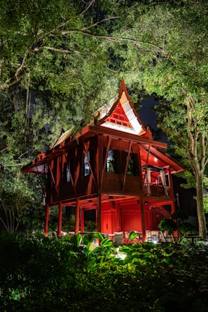 An elevated traditional wooden house with a steeply pitched roof is illuminated at night. It is surrounded by lush greenery, with tall trees and various plants visible in the foreground. The house is painted red, with light highlighting its architectural details and casting shadows in the surrounding foliage.