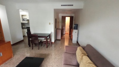 A living space with a brown couch and beige cushions on the right side of the room. A small dining table with four chairs is placed near the center, with an open kitchen visible in the background. The room has marbled flooring, and there is a water dispenser next to the entrance leading to another room.