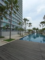 A modern high-rise building with glass windows stands alongside a long, rectangular swimming pool. Palm trees and neatly landscaped greenery line the walkway, creating a serene and upscale environment. A wooden deck area is visible in the foreground, and the sky is overcast.