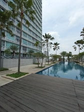 A modern high-rise building with glass windows stands alongside a long, rectangular swimming pool. Palm trees and neatly landscaped greenery line the walkway, creating a serene and upscale environment. A wooden deck area is visible in the foreground, and the sky is overcast.