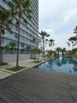 A modern high-rise building with glass windows stands alongside a long, rectangular swimming pool. Palm trees and neatly landscaped greenery line the walkway, creating a serene and upscale environment. A wooden deck area is visible in the foreground, and the sky is overcast.