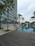 A modern high-rise building with glass windows stands alongside a long, rectangular swimming pool. Palm trees and neatly landscaped greenery line the walkway, creating a serene and upscale environment. A wooden deck area is visible in the foreground, and the sky is overcast.