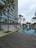 A modern high-rise building with glass windows stands alongside a long, rectangular swimming pool. Palm trees and neatly landscaped greenery line the walkway, creating a serene and upscale environment. A wooden deck area is visible in the foreground, and the sky is overcast.