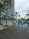 A modern high-rise building with glass windows stands alongside a long, rectangular swimming pool. Palm trees and neatly landscaped greenery line the walkway, creating a serene and upscale environment. A wooden deck area is visible in the foreground, and the sky is overcast.