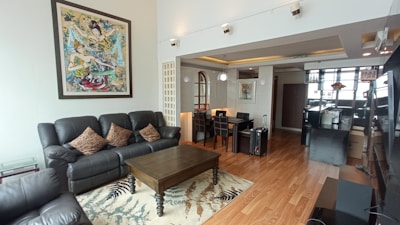 A modern living room with a large black leather sofa set and brown cushions, centered around a dark wooden coffee table on a patterned rug. The room features wooden flooring and is well-lit with natural light from large windows. On the wall, there's a colorful abstract painting. The dining area includes a dark dining table with high-back chairs under a decorative suspended light. Various decorative items and packed boxes are visible, indicating a contemporary and somewhat transitional space.