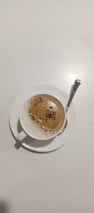 A cup of frothy coffee is placed on a white saucer with a silver spoon resting on the saucer. The coffee has a creamy top layer with specks of cocoa or cinnamon sprinkled on it.