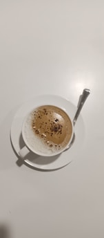 A cup of frothy coffee is placed on a white saucer with a silver spoon resting on the saucer. The coffee has a creamy top layer with specks of cocoa or cinnamon sprinkled on it.