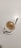 A cup of frothy coffee is placed on a white saucer with a silver spoon resting on the saucer. The coffee has a creamy top layer with specks of cocoa or cinnamon sprinkled on it.