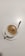A cup of frothy coffee is placed on a white saucer with a silver spoon resting on the saucer. The coffee has a creamy top layer with specks of cocoa or cinnamon sprinkled on it.