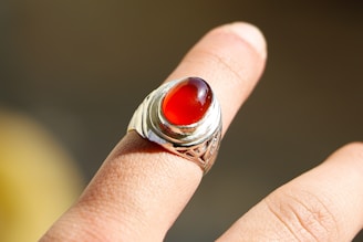 A silver ring featuring a large, oval red gemstone is worn on an adult's finger. The background is blurred, highlighting the intricacy of the ring's design and the vividness of the gemstone.