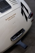 A close-up view of the rear section of a classic Porsche Carrera, featuring its distinctive emblem in gold lettering. The image showcases the car's sleek, curved body design with a vented engine cover and tail lights. Large black stripes add a dynamic element to the cream-colored exterior.