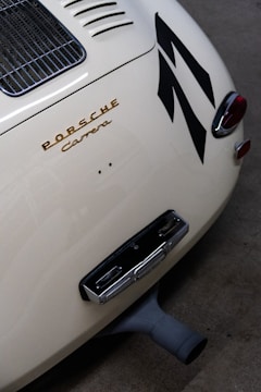 A close-up view of the rear section of a classic Porsche Carrera, featuring its distinctive emblem in gold lettering. The image showcases the car's sleek, curved body design with a vented engine cover and tail lights. Large black stripes add a dynamic element to the cream-colored exterior.