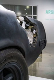 The rear of a matte black car reveals an artistic detail where the body appears to be eroded, exposing a cluster of crystals and minerals inside. This contrast between the rough exterior and the delicate, shimmering interior creates an intriguing visual impact.