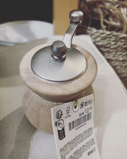 A close-up view of a manual coffee grinder with a wooden body and metallic top. A price tag is attached, displaying EUR 12,99 along with other product details and barcodes. The background includes a woven basket and a light-colored fabric, possibly indicating a rustic or domestic setting.
