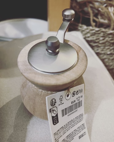 A close-up view of a manual coffee grinder with a wooden body and metallic top. A price tag is attached, displaying EUR 12,99 along with other product details and barcodes. The background includes a woven basket and a light-colored fabric, possibly indicating a rustic or domestic setting.
