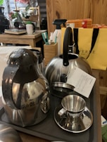 A collection of kitchenware items including a stainless steel kettle, a thermal carafe, and a metallic cup with a small saucer. They are placed on a dark tray atop a wooden table. In the background, there is a yellow shopping bag, a box of tea, various kitchen utensils, and other household items.