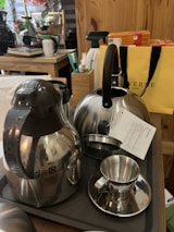 A collection of kitchenware items including a stainless steel kettle, a thermal carafe, and a metallic cup with a small saucer. They are placed on a dark tray atop a wooden table. In the background, there is a yellow shopping bag, a box of tea, various kitchen utensils, and other household items.