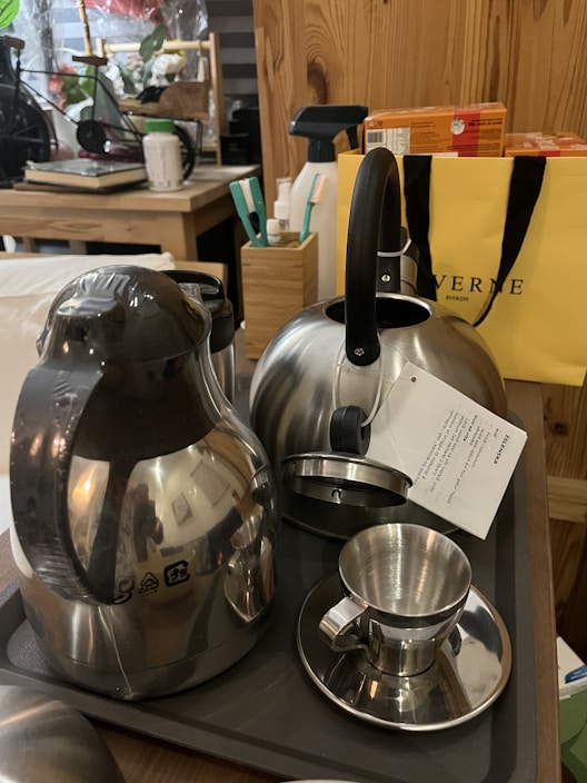 A collection of kitchenware items including a stainless steel kettle, a thermal carafe, and a metallic cup with a small saucer. They are placed on a dark tray atop a wooden table. In the background, there is a yellow shopping bag, a box of tea, various kitchen utensils, and other household items.