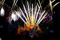 A grand castle illuminated at night with several fireworks exploding in the sky above it, creating a vibrant and magical atmosphere. The fireworks display includes various colors and patterns, complementing the illuminated castle.