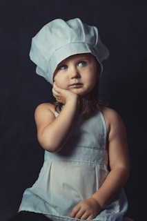 A young child wearing a white chef's hat and apron appears deep in thought, with one hand resting on their cheek. The background is dark, which highlights the child’s facial expression and clothing.