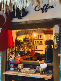 A small coffee shop window adorned with decorative elements, including a tribal mask and a colorful bird sculpture. There's a Turkish flag hanging to the left. The counter displays a basket with paper cups and sugar packets, and a coffee grinder is visible inside.
