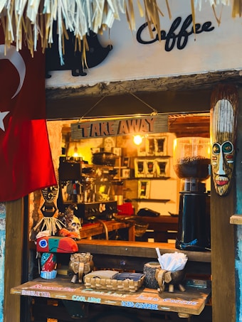 A small coffee shop window adorned with decorative elements, including a tribal mask and a colorful bird sculpture. There's a Turkish flag hanging to the left. The counter displays a basket with paper cups and sugar packets, and a coffee grinder is visible inside.