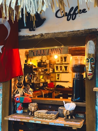 A small coffee shop window adorned with decorative elements, including a tribal mask and a colorful bird sculpture. There's a Turkish flag hanging to the left. The counter displays a basket with paper cups and sugar packets, and a coffee grinder is visible inside.