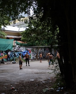 Several shirtless individuals are gathered in an urban outdoor area that appears to be an improvised sports court. The setting is surrounded by trees and a wall covered in colorful graffiti. In the background, there are some buildings partially visible. The ground is littered with some debris and leaves.