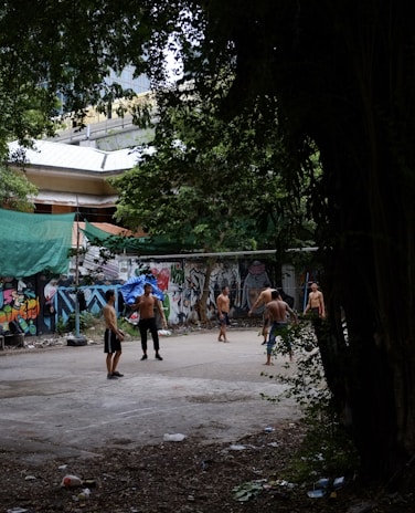 Several shirtless individuals are gathered in an urban outdoor area that appears to be an improvised sports court. The setting is surrounded by trees and a wall covered in colorful graffiti. In the background, there are some buildings partially visible. The ground is littered with some debris and leaves.