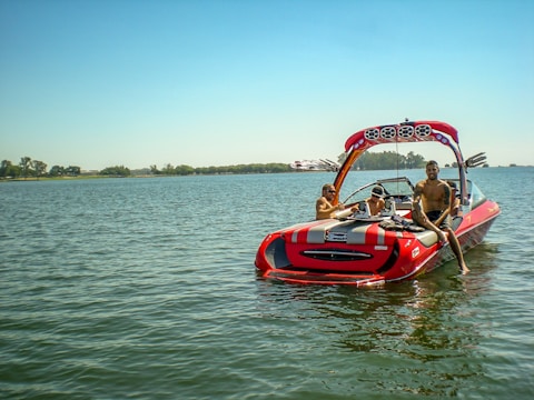 A red and gray speedboat is on a calm lake with three people onboard. The boat features a large wakeboard tower with speakers, and the individuals appear relaxed, enjoying the sunny weather. Trees line the distant shore under a clear blue sky.
