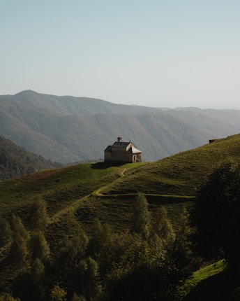 A solitary house sits atop a lush green hill, surrounded by a vast expanse of rolling mountains under a clear sky. A narrow path leads to the house, flanked by various trees and dense foliage.
