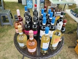 A round wicker table is set up outdoors, displaying a variety of wine bottles. There are different types of wine, such as red, white, and rosé, with colorful labels featuring various designs. The surroundings include grass and nearby plastic chairs, along with scattered outdoor items suggesting a picnic or sale event.
