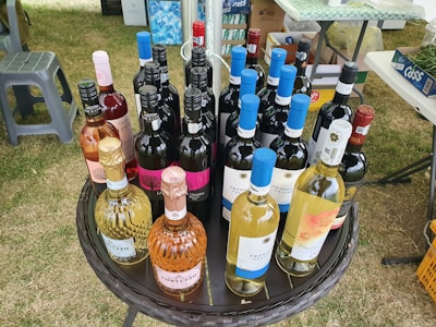 A round wicker table is set up outdoors, displaying a variety of wine bottles. There are different types of wine, such as red, white, and rosé, with colorful labels featuring various designs. The surroundings include grass and nearby plastic chairs, along with scattered outdoor items suggesting a picnic or sale event.