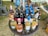 A round wicker table is set up outdoors, displaying a variety of wine bottles. There are different types of wine, such as red, white, and rosé, with colorful labels featuring various designs. The surroundings include grass and nearby plastic chairs, along with scattered outdoor items suggesting a picnic or sale event.