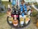 A round wicker table is set up outdoors, displaying a variety of wine bottles. There are different types of wine, such as red, white, and rosé, with colorful labels featuring various designs. The surroundings include grass and nearby plastic chairs, along with scattered outdoor items suggesting a picnic or sale event.