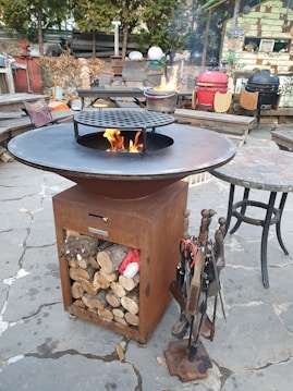 A rust-colored outdoor fire pit with a circular metal grate and visible flames. It is placed on a stone patio and surrounded by various barbecue grills. Wood logs are stored in the base compartment of the fire pit. Various grill tools are hanging on the side. The setting is rustic with greenery and eclectic decorations in the background.