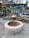 A cozy outdoor setting featuring a fire pit in the center, surrounded by a stone patio. There is a stack of firewood next to a wooden structure adorned with string lights and greenery. A chair is placed nearby, creating a warm and inviting atmosphere.