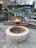 A cozy outdoor setting featuring a fire pit in the center, surrounded by a stone patio. There is a stack of firewood next to a wooden structure adorned with string lights and greenery. A chair is placed nearby, creating a warm and inviting atmosphere.